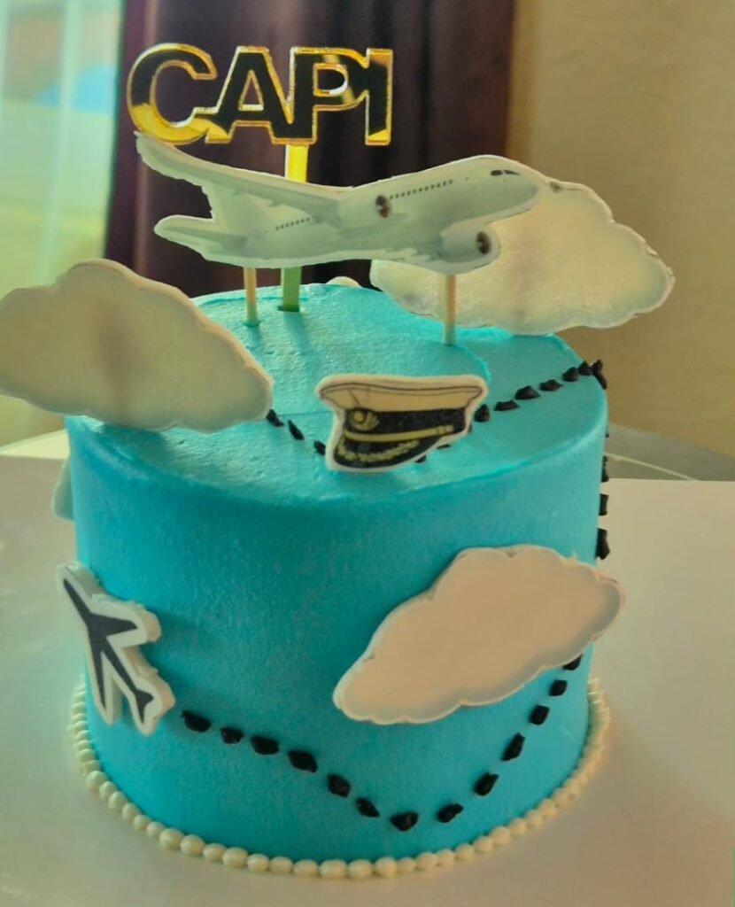 Pastel azul con topper de avión, nubes y la palabra Capi marcando una ruta de piloto