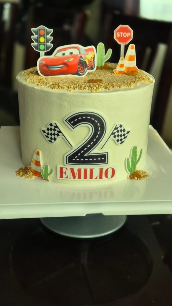 Pastel infantil personalizado temático de Cars con el número 2, nombre Emilio y decoraciones de carreteras, conos y Rayo McQueen.