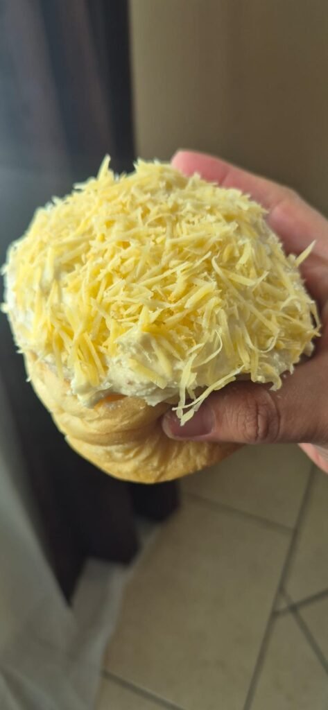 Rol de canela cubierto con betún y queso rallado de bola sostenido en la mano, con textura suave y esponjosa.