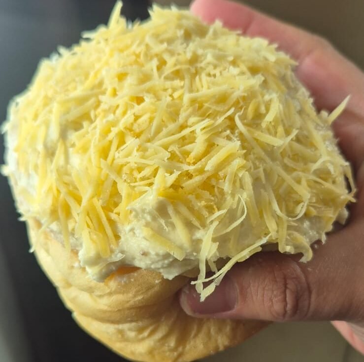 Rol de canela cubierto con betún y queso rallado de bola sostenido en la mano, con textura suave y esponjosa.